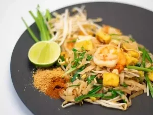 Gebratene Reisnudeln mit Garnelen, Tofu, Eiern, Mungbohnen-Sprosse und Schnittknoblauch, serviert mit Limetten, Chilipulver und gerösteten Erdnüssen auf einem schwarzen Teller.